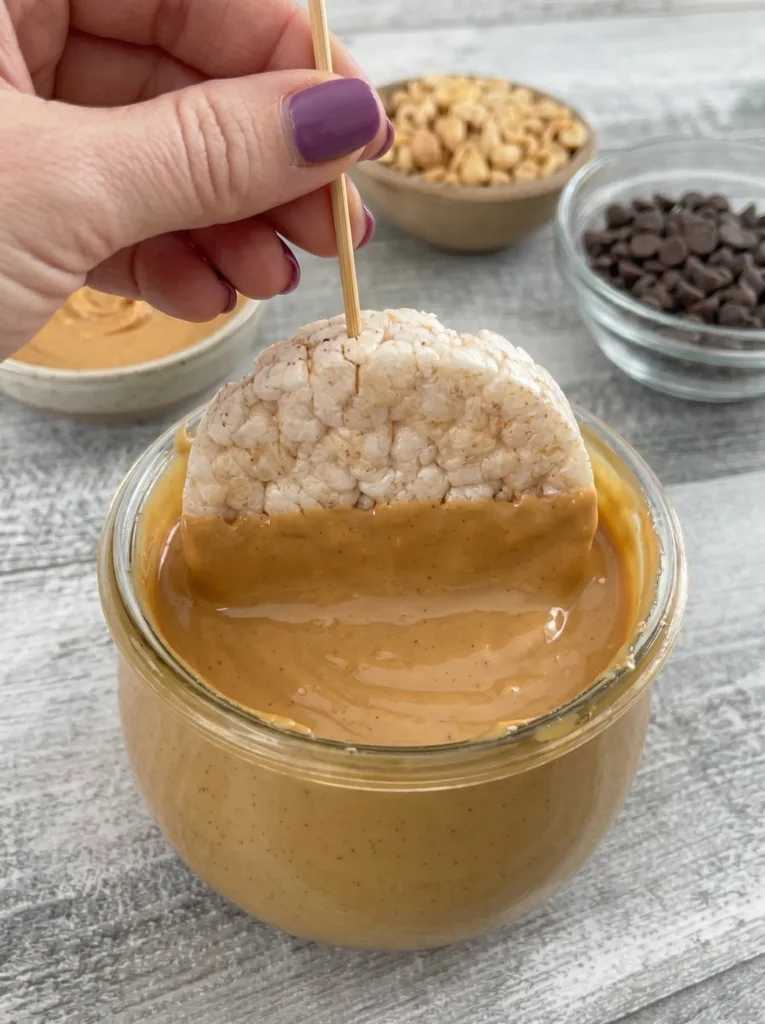 Rice cake on a skewer being dipped face-down into a jar of peanut butter.